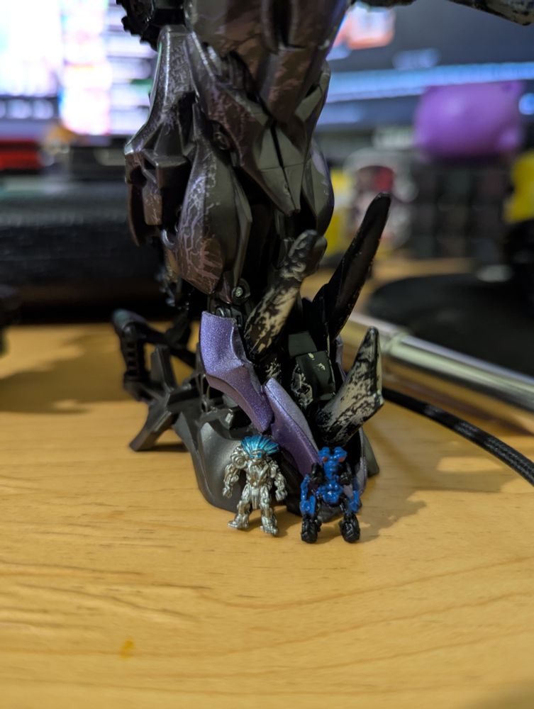 A picture of the actual toys from the box in the previous picture zoomed in to focus on the smaller figures. Only Shockwave's foot can be seen and the smaller figures are little more than indistinct shapes that are barely taller than the toe of the foot they're stood in front of.