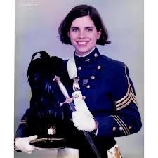 Photograph of a young Nancy Mace circa 1999 in full-dress uniform from The Citadel Military College of South Carolina. She is wearing a blue wool jacket with 3 rows of brass buttons, although only 1 row is visible. Her sleeves have 3 stripes, indicating that she has achieved the rank of C4, diagonally, across her chest is a white belt with brass breastplate, and she is holding her white-handled Cadet Officer's sword in her left hand [right side of the picture]. In her right hand she is holding a black shako [a tall, cylindrical military cap, with a visor], and because she is senior with rank, her shako has a plume.

She is smiling broadly, with her brown, jaw-length hair tucked behind her ears.
