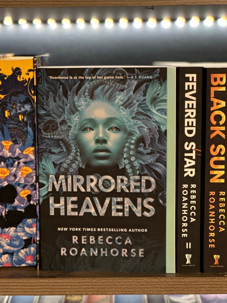 This photo shows the paperback edition of Mirrored Heavens by Rebecca Roanhorse on a wooden bookstore shelf, nestled between other books in the Between Earth and Sky series. The cover art features an image of a woman’s face in cool shades of teal and blue. Her eyes glow faintly, and her hair is an intricate mass of feathers, braids, and sea creature-like shapes. Above her head is a endorsement from R.F. Kuang that reads: “Roanhorse is at the top of her game here.”