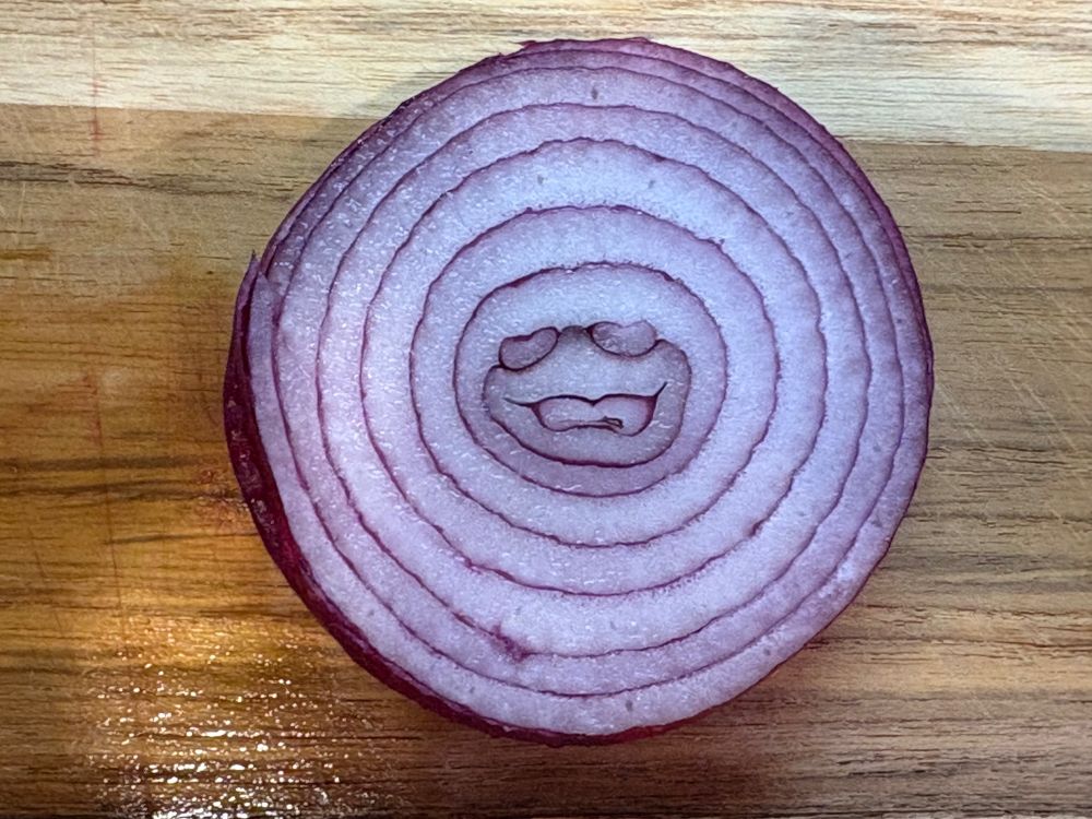 A slice of red Bermuda onion, on a cutting board, with what looks like the face of a stoned person in the middle of the onion's concentric rings.