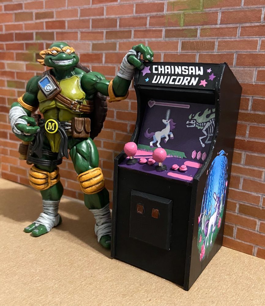 Action figure of the ninja turtle Michaelangelo standing next to the Chainsaw Unicorn arcade game, resting one hand on the top of the machine.