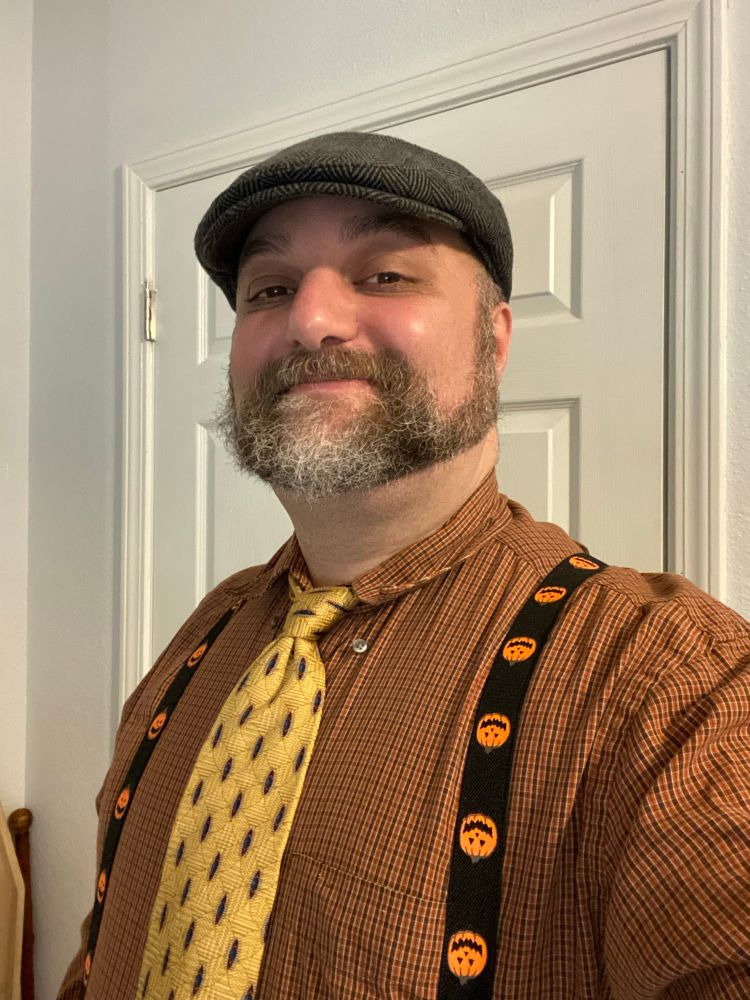 Myself (Caucasian man in his forties, salt-and-pepper short beard) in Halloween clothes: an orange plaid button-up, yellow tie, tweed cap, and suspenders with Jack-O-Lanterns.