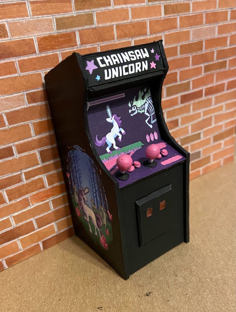 1:18 scale arcade game. Marquee reads “Chainsaw Unicorn,” with stars around it. On the side, a unicorn with a chainsaw for a horn faces off against zombie unicorns. Joysticks and buttons are pink. The cabinet features two transparent red coin slots.