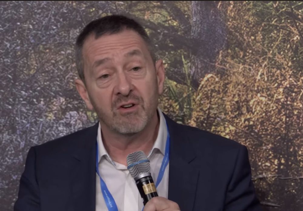 Chris Boardman speaking into a microphone at COP29 in front of a backdrop of foliage