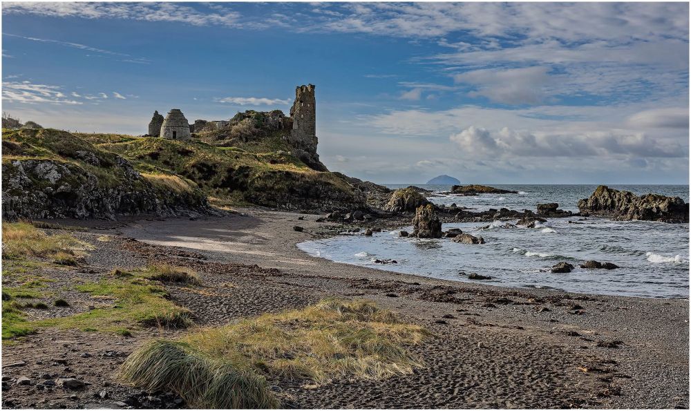 Image of a coastal landscape featuring an ancient ruin atop a grassy hillside, with a sandy beach and rocky shoreline in the foreground. The sky is partly cloudy, and an island is visible in the distance.