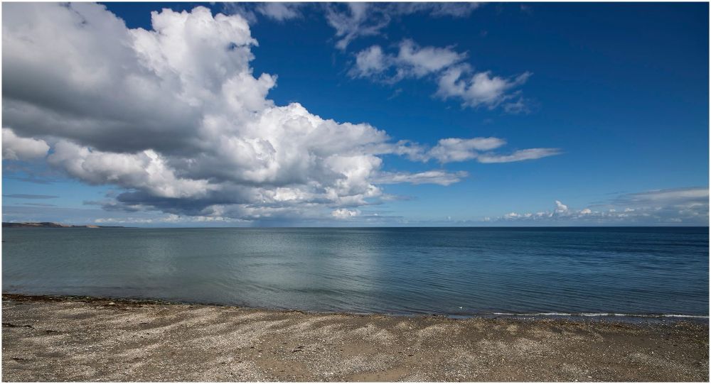 A serene coastal scene featuring a calm sea under a bright blue sky with dramatic white clouds. The sandy shore is visible in the foreground, with gentle waves lapping at the beach.