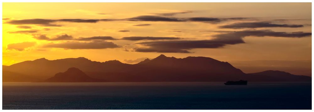 A silhouette of mountains under a golden sunset, with a calm sea in the foreground and a ship in the distance.