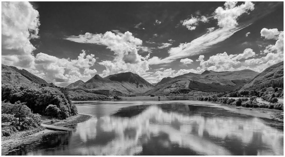 A black and white landscape featuring a serene lake surrounded by mountains and lush greenery. Fluffy clouds reflect in the calm water, creating a picturesque and tranquil scene.