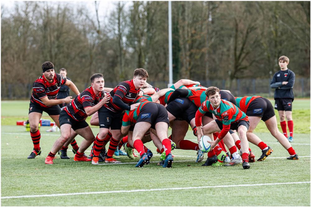 A rugby match in progress on a grass field. Players in red and black jerseys are engaged in a scrum, while one player in a green and red jersey prepares to pass the ball. Other players are observing the play.