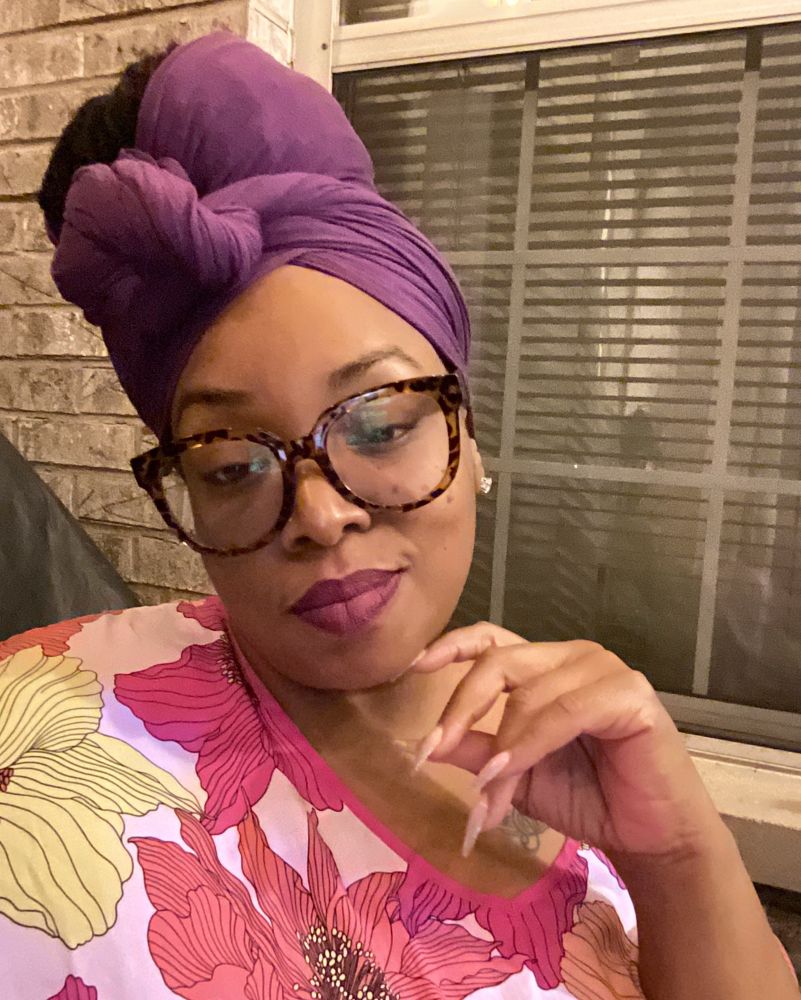  Black woman in purple head wrap with locs in a bun. Wearing a military colored mumu 