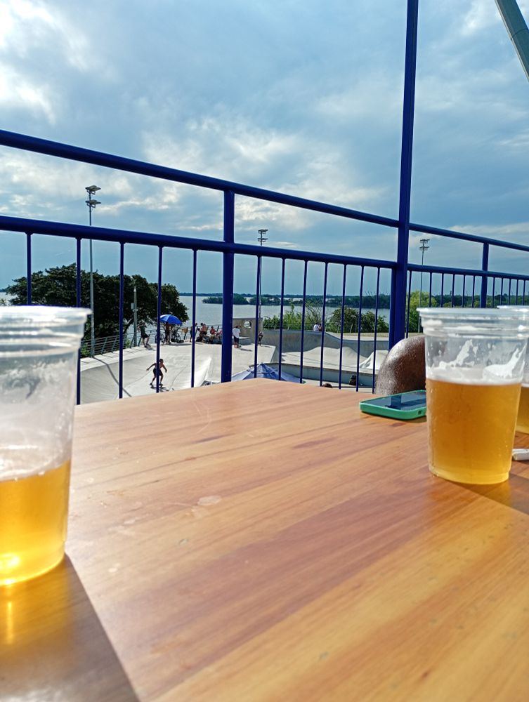 Foto diurna, dois copos de cerveja numa mesa de madeira e ao fundo uma pista de skate e o rio Guaíba 