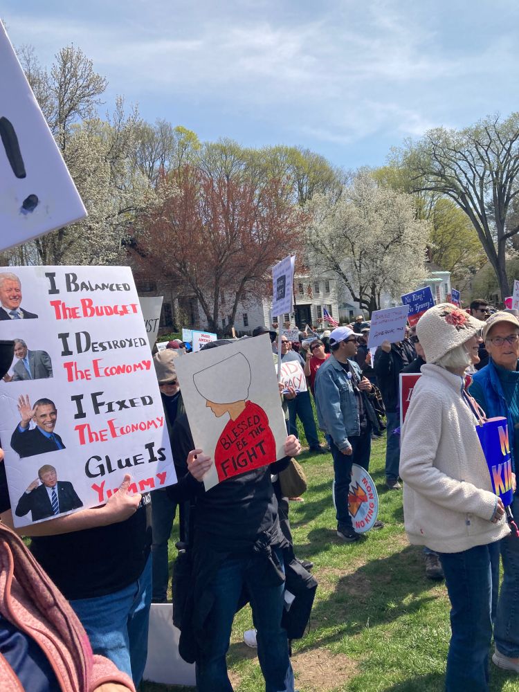 General crowd photo with two visible signs. One has photos of four presidents, with financial text. Clinton: I balanced the budget. Bush: I destroyed the economy. Obama: I fixed the economy. Trump: Glue is Yummy. Next to the person holding that sign is a woman holding one that has a profile drawing of a "handmaiden" in white cap and red garment, which reads "Blessed is the Fight."