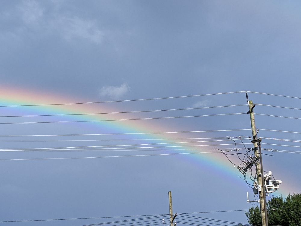 A bright rainbow arc intersects a set of transformers on a power pole.