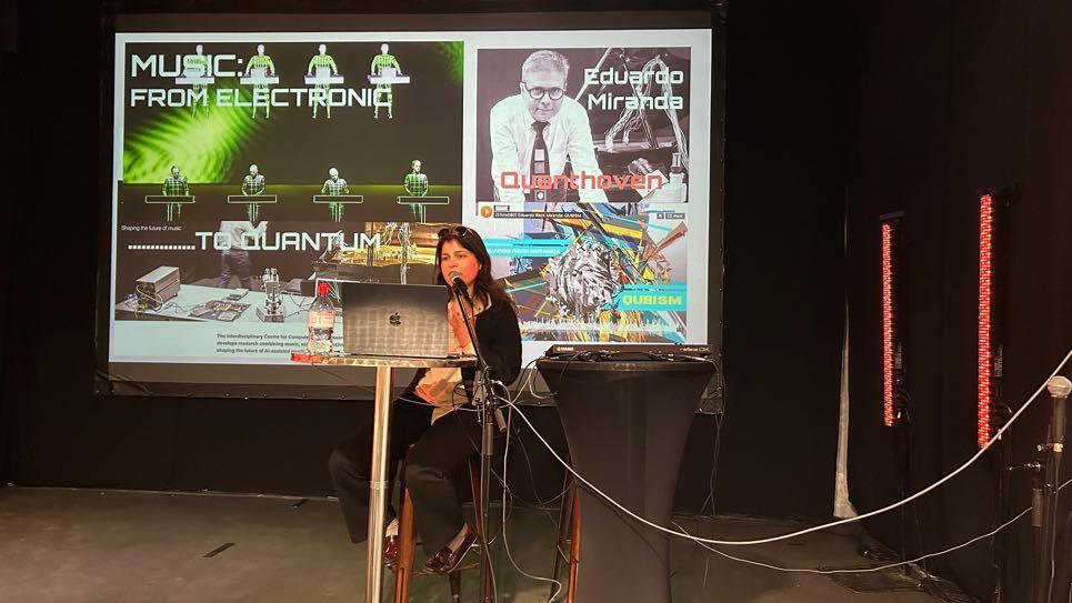 A woman sat behind a laptop presenting on a big screen. The screen reads: 'Music from electronic to quantum; Eduardo Miranda'