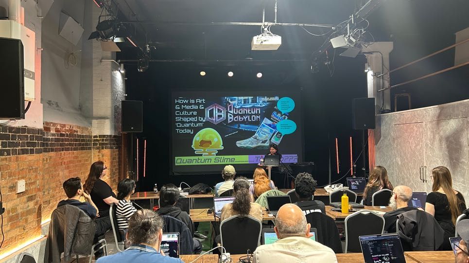 A woman sat behind a laptop presenting on a big screen. The screen reads: 'How is art and media and culture shaped by quantum today'