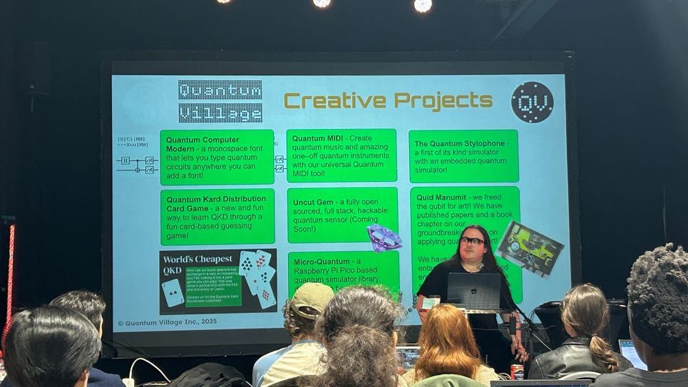 A man sat behind a laptop presenting on a big screen. The screen reads: 'Quantum Village creative projects'