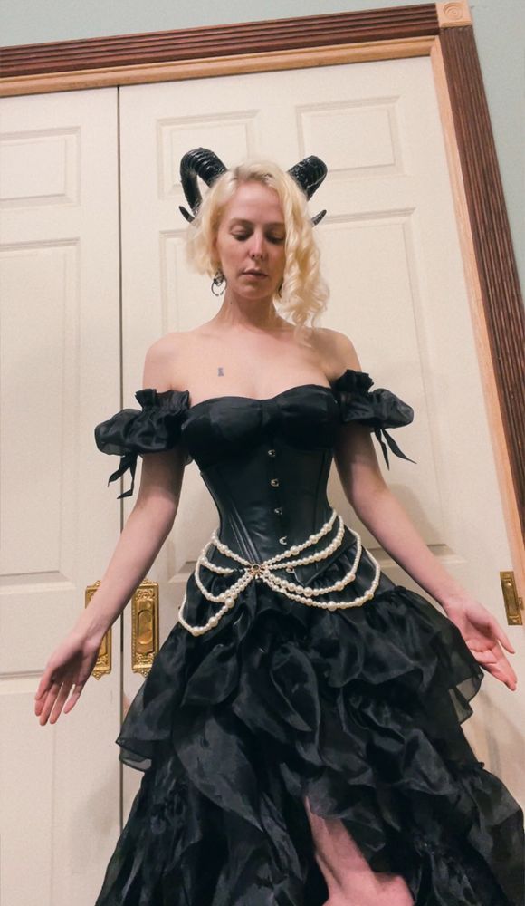 A glossy black gown with a high slit in the (very voluminous) skirt, puffy sleeves (shown worn off the shoulder) and a black leather corset worn over it with faux pearls draped around the waist.