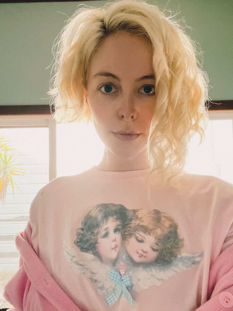 A backlit selfie of a white girl with blonde curly hair cut into asymmetrical shaggy bob, wearing a pastel pink tshirt with two cherubs (also with fluffy ringlets), and a slightly-darker-pink fuzzy onesie worn off-the-shoulder.