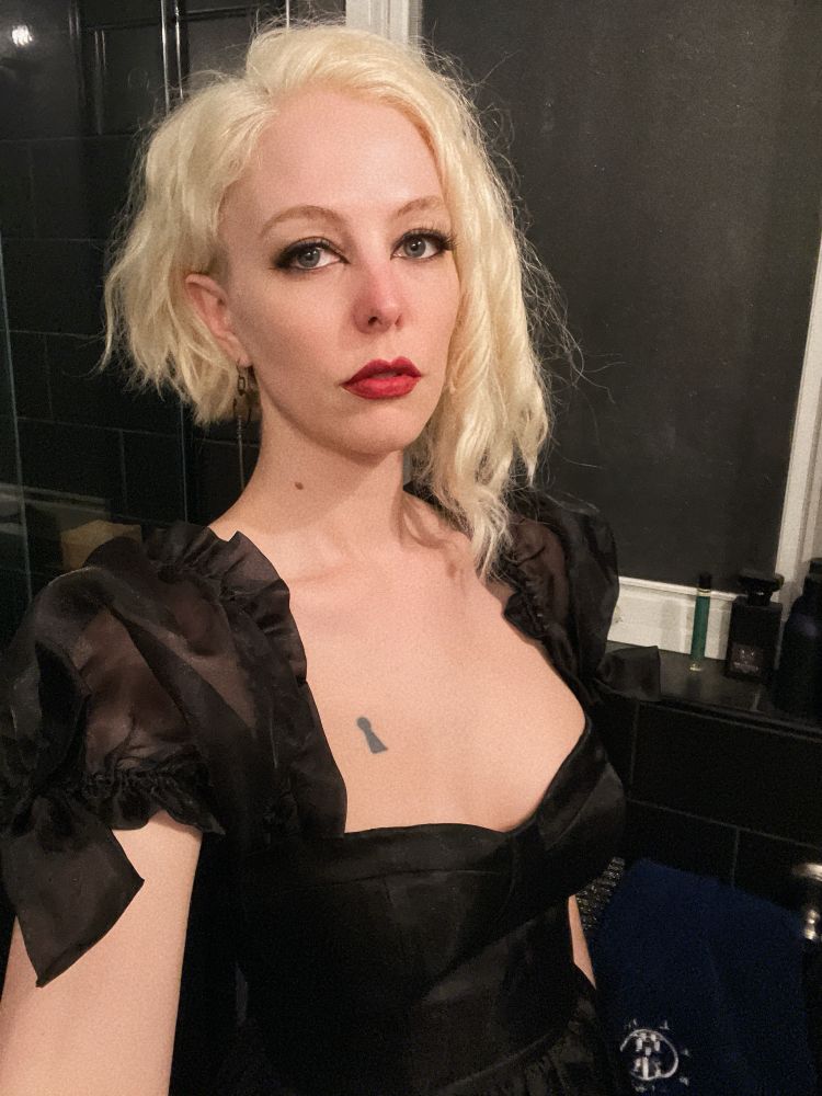 Bathroom selfie of a tall slender white girl with a shaggy platinum blonde asymmetrical bob, and bold makeup. Wearing a fluffy black ballgown.