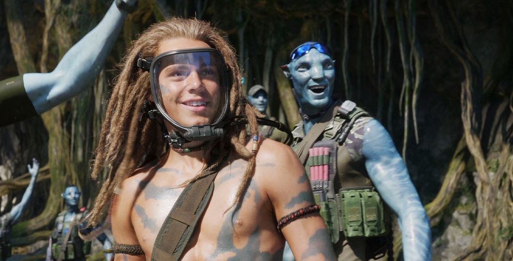 A shot from AVATAR: THE WAY OF WATER. Spider, the dreadlocked human born on Pandora, is wearing his breather and smiling while standing in front of smiling Na'vi super soldiers.