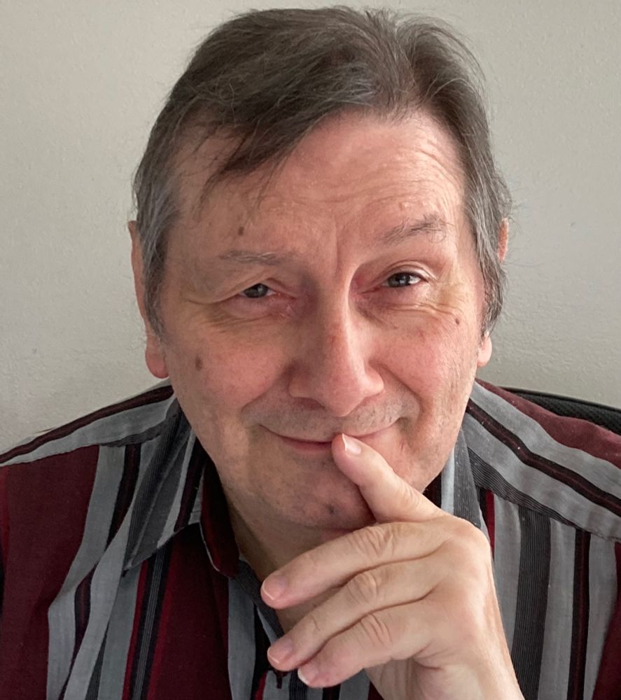 A handsome fellow with dark hair and hazel eyes wearing a striped shirt. He is staring at the camera, wearing a mischievous grin, and has one hand on his chin.