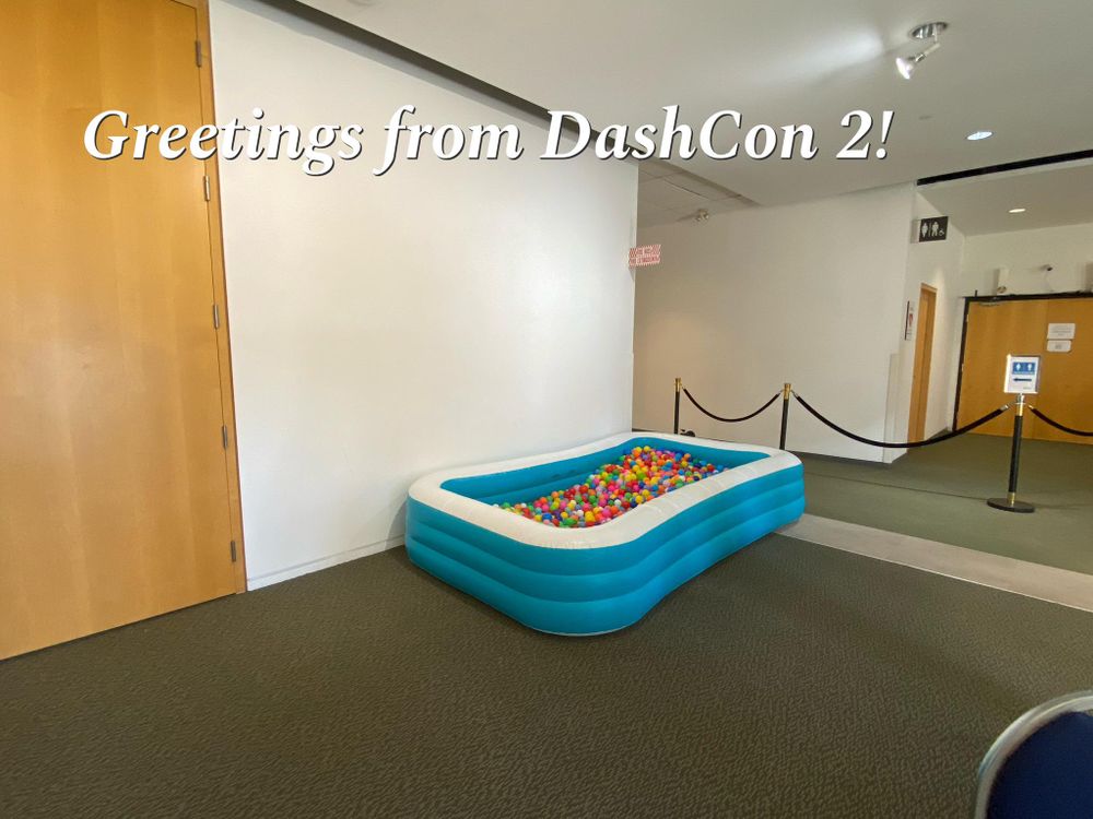 Fotografía de un espacio liminal, paredes blancas, unas puertas marrón claro al fondo, una moqueta gris, con una piscina hinchable en el medio llena de bolas de plástico.
Sobre la imagen en texto con serifa blanco está escrito "Greetings from DashCon 2!"