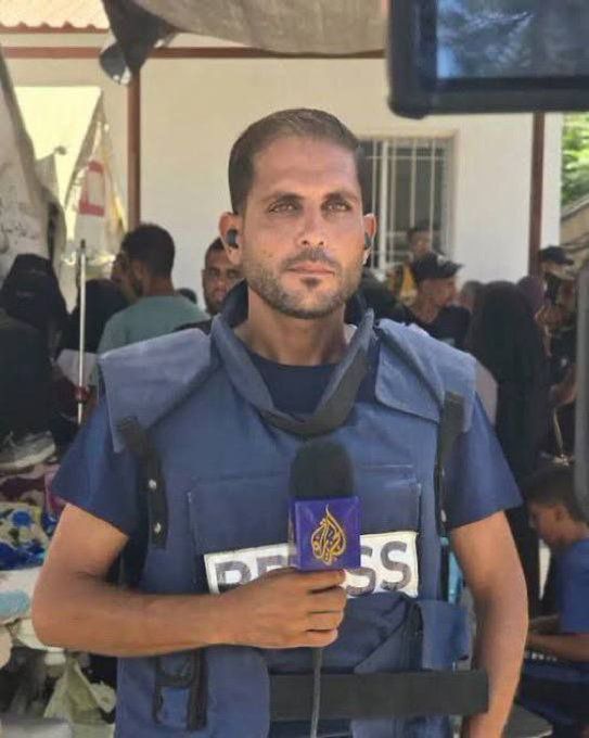 a man holds a microphone with an al jazeera logo while wearing a vest with the word press on it