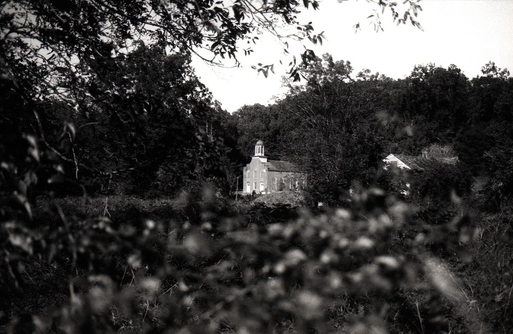 a photo of a woody area shows an old church in the distance, surrounded by woods and trees. Photo by Ashton Pittman