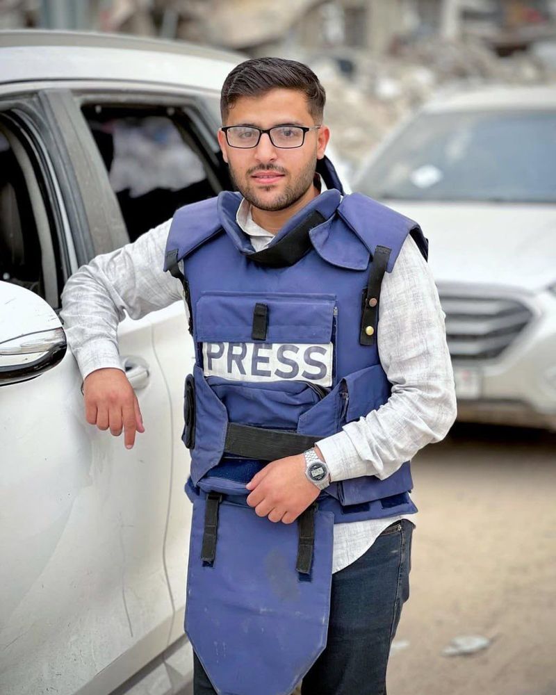 a young man in glasses wearing a protective vest with the word press on it leans against a car 