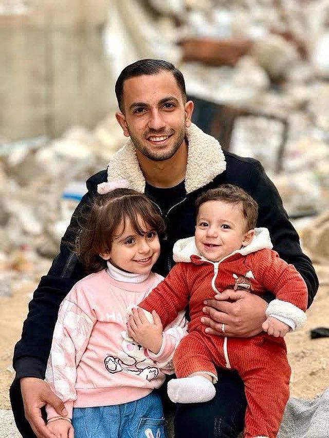 a young man holds a young boy and girl in front of rubble 