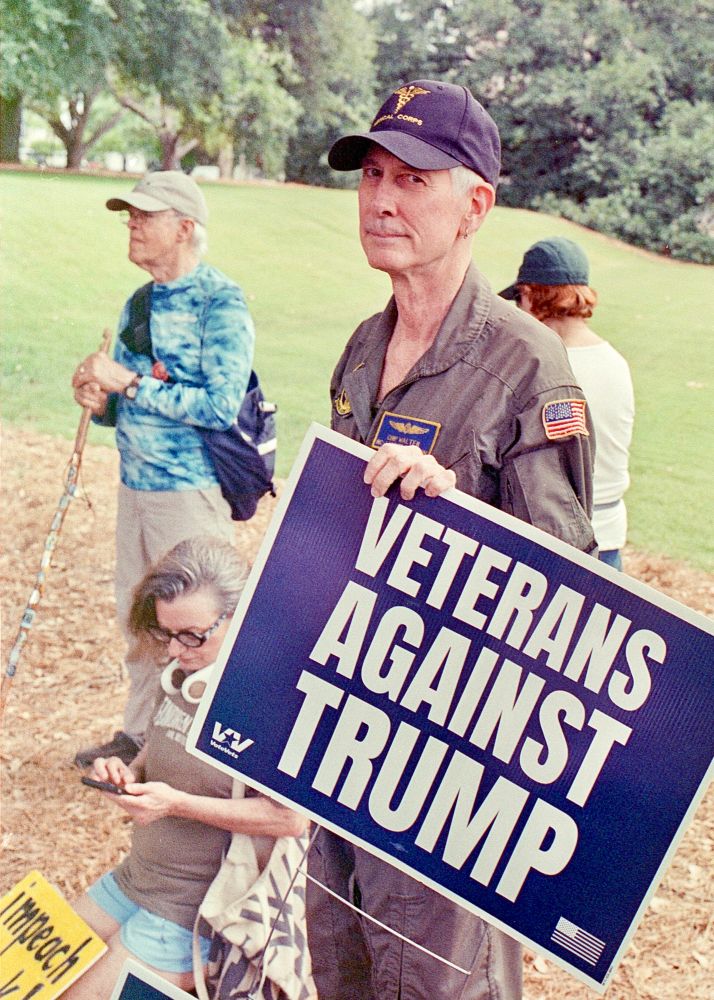 a photo of a man who appears to be in his 60s or 70s is wearing a blue cap that says "medical corps" and holding a sign that says "veterans against trump" with several other protesters behind him standing on the grass 