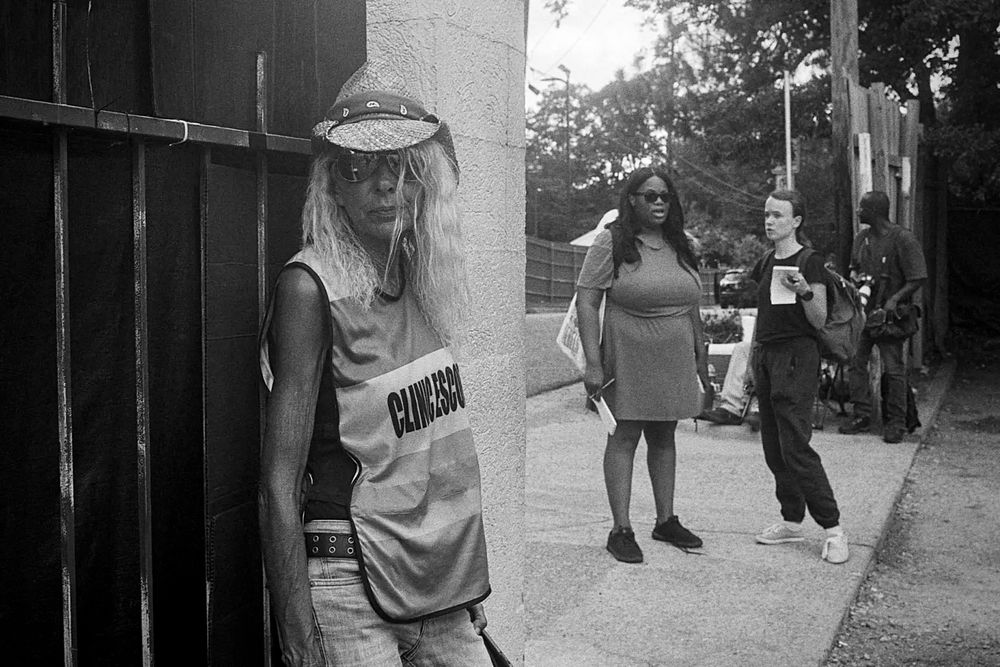 a grainy black and white photo shows a woman with chest-length blond hair, sunglasses and a straw hat wearing a vest that says "CLINIC ESCORT" on it and leaning against a black wrought iron fence, while two women stand in a driveway behind her speaking 