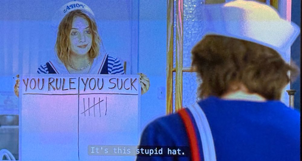 A frame from the third season of “Stranger Things.” Robin (Maya Hawke) and Steve (Joe Keery) are both wearing the cheesy sailor suits required for employees of Scoops Ahoy. She confronts him with a whiteboard chart tallying his successes (“YOU RULE”) and failures (“YOU SUCK”). The former shows no marks; the latter, six. The subtitle reads, “It’s this stupid hat.” 