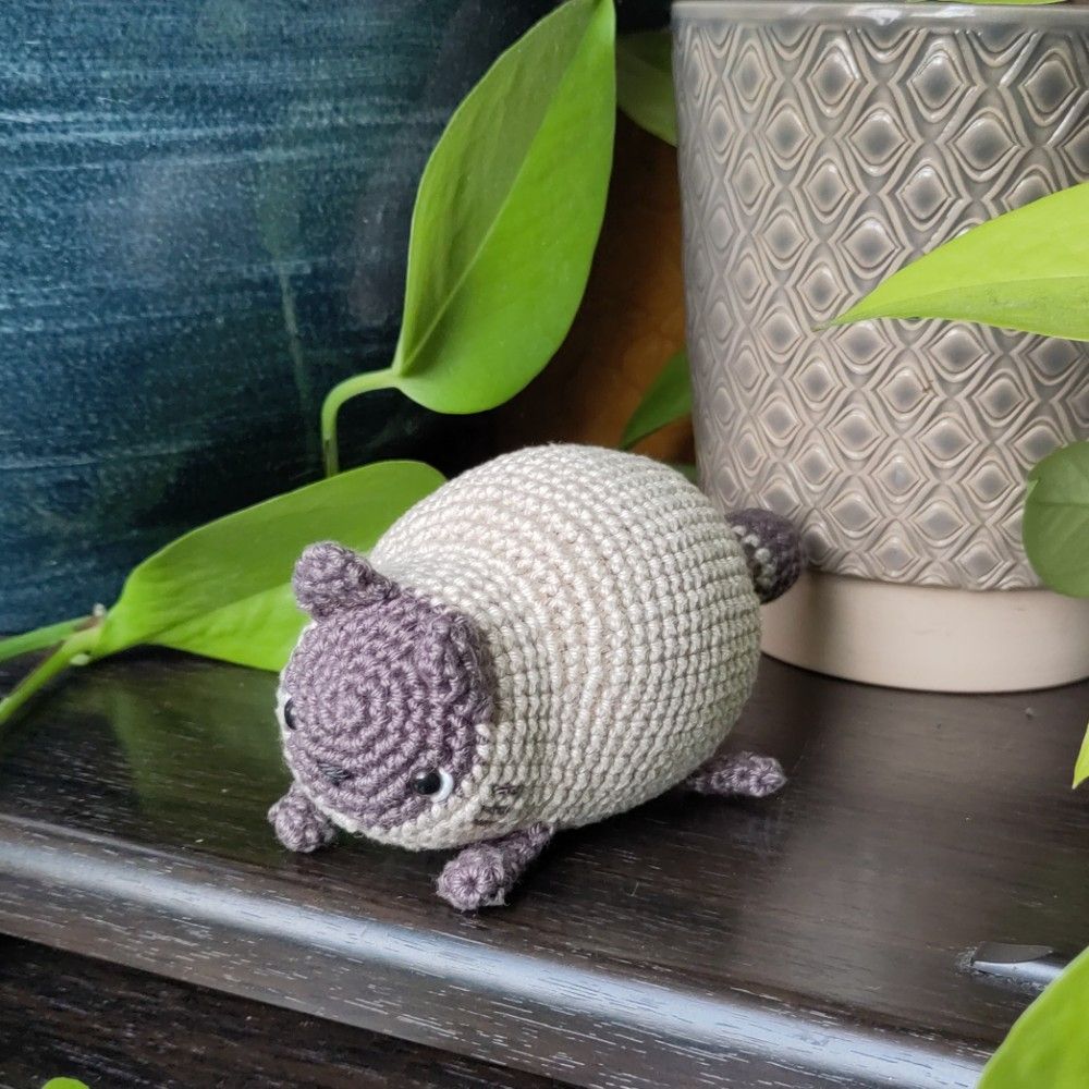 A chonky crocheted siamese cat lies on a wooden surface in front of potted plants. 
The cat is beige with reddish-brown points.