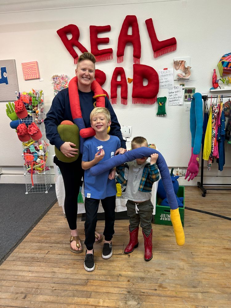 Family of three holding up silly plush toys shaped like olives and multicolor tubes. 