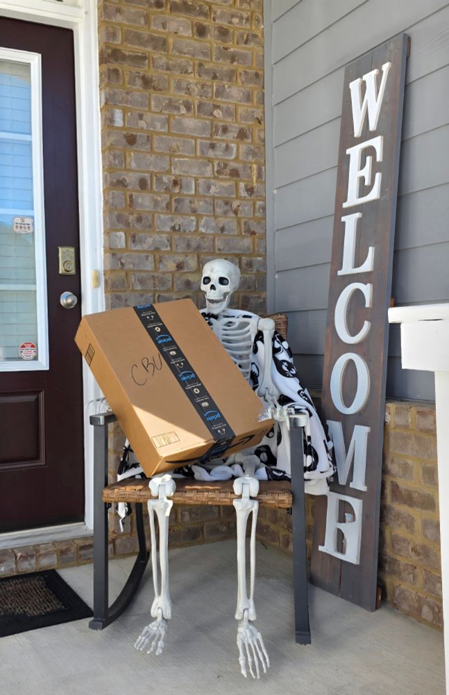 A skeleton, Halloween decoration is sitting in a rocking chair on a front porch with an Amazon package in its lap. (A welcome sign is behind them)