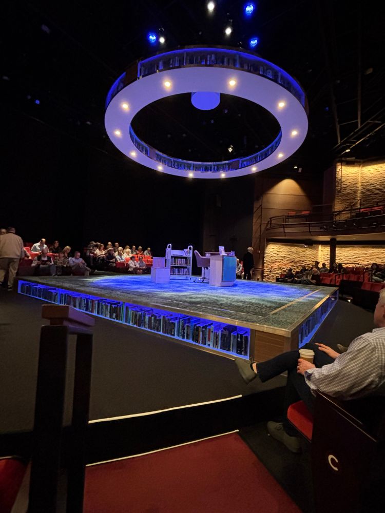 Photo of the stage of a play. The base is bordered with books (all banned books). Suspended from the ceiling is a large circle ring and in the ring are all banned books too.