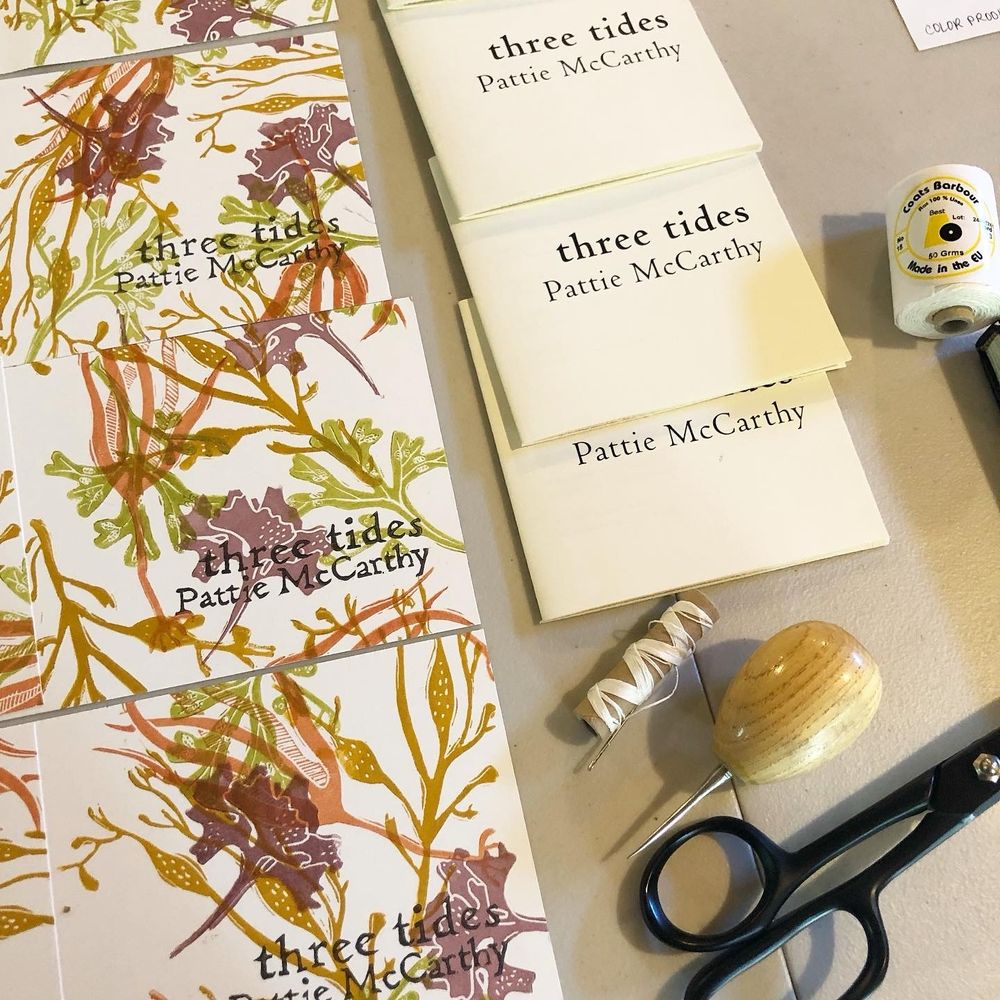 Process shot of trimmed covers for “three tides” by Pattie McCarthy alongside some folded and trimmed inner booklets, binding thread and tools. The cover features a multiblock linocut pattern of seaweeds and hand-carved lettering. Design © Shanna Compton for Bloof Books, 2022.