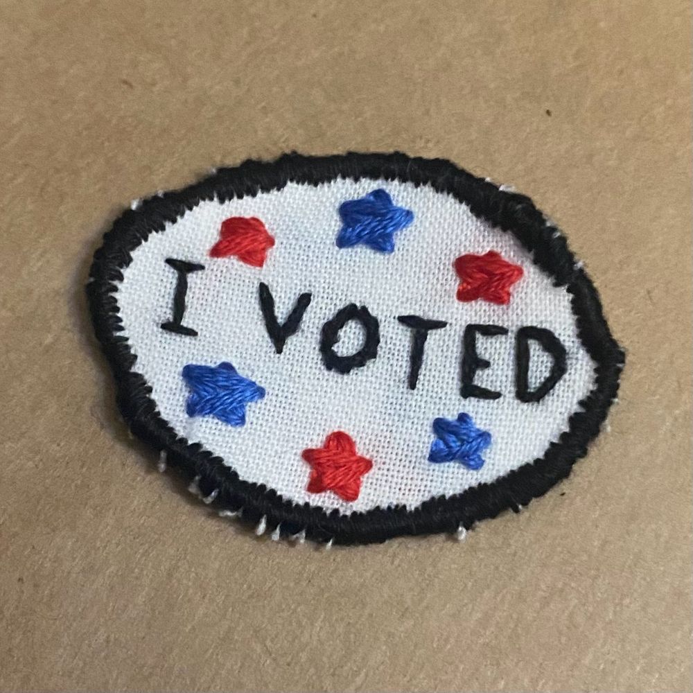 A hand-sewn oval patch. White fabric, uneven black outline, and the words “I VOTED” stitched in black and surrounded by red and blue stars.
