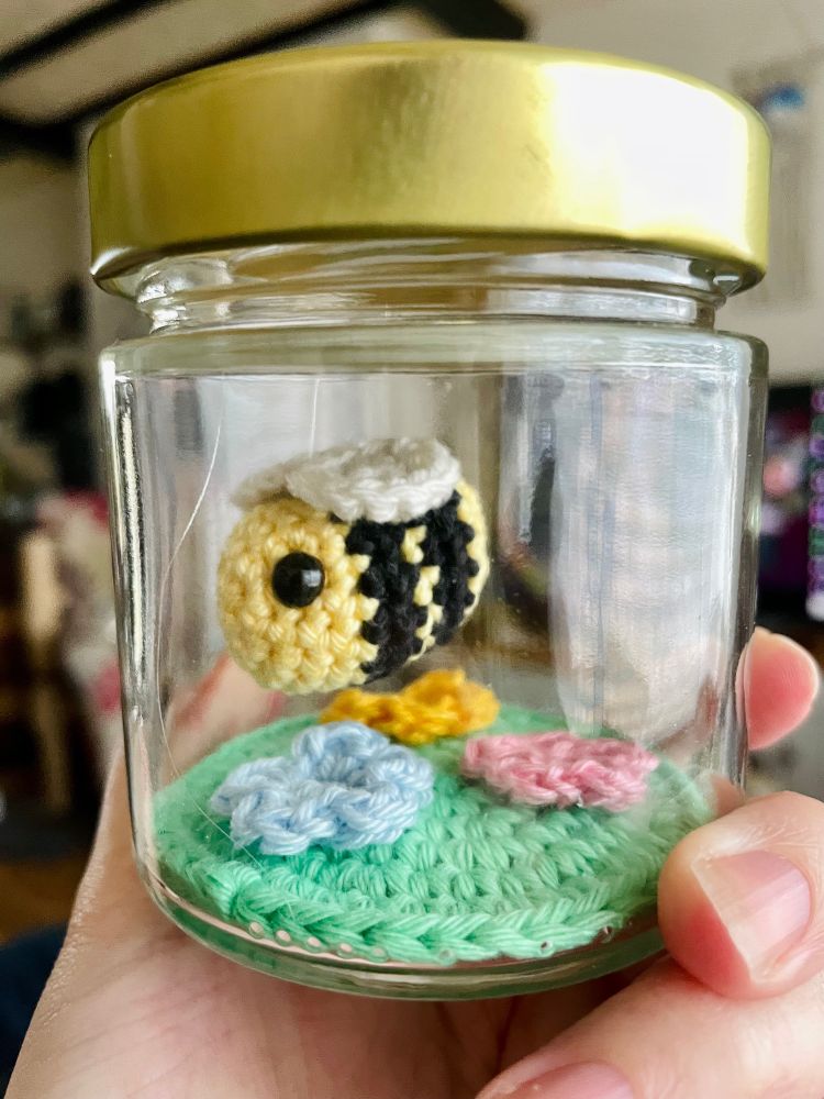a small jar with a crochet bee hanging inside, and a small patch of crochet grass and flowers on the bottom.