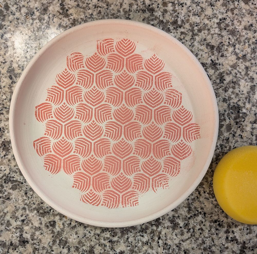 Bisqueware plate on counter dwcoraed with underglaze transfer pattern. There is a round sponge in the bottom right corner of the frame. 
