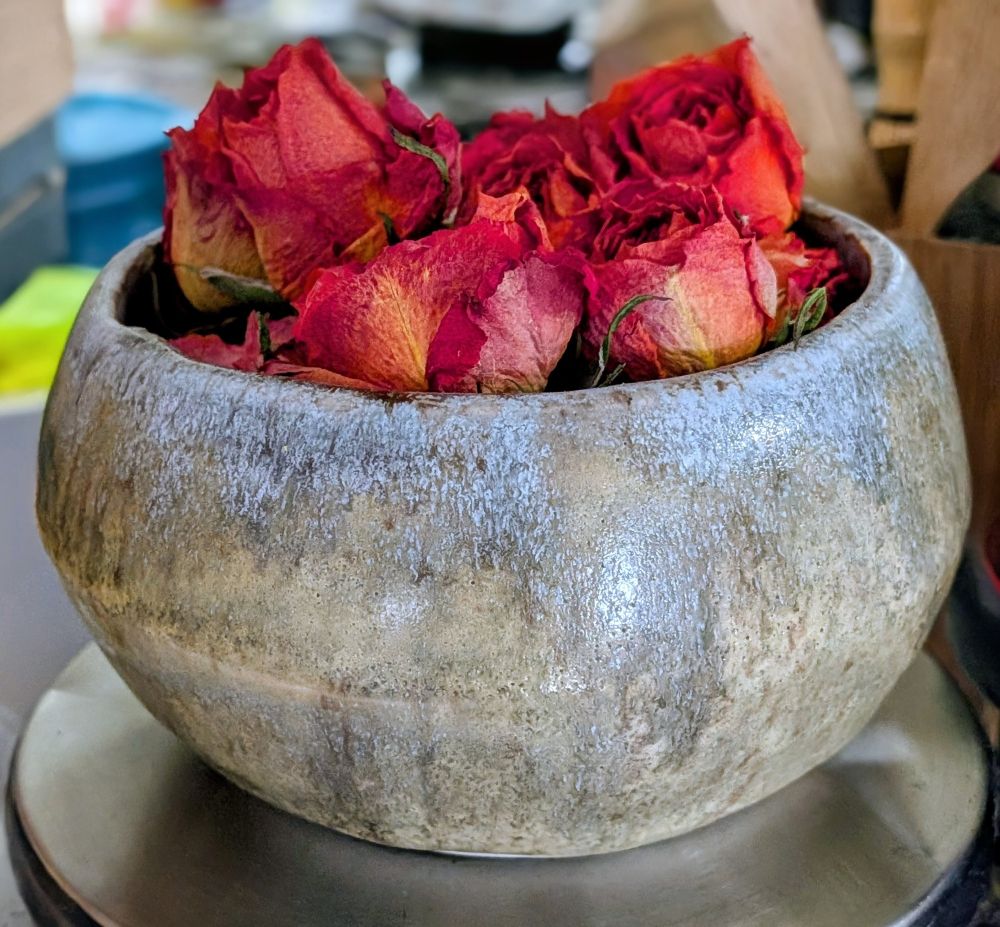 A small grey ceramic bowl containing a bunch of small red dry rose buds sitting on a pedestal.