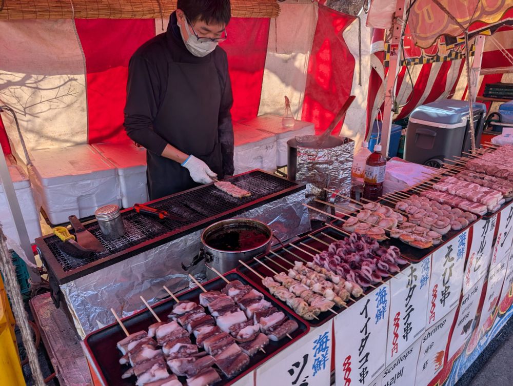Person in mask grills pork belly on stick atop a grill.  Behind him is the back of his booth tent.  In front of him is a spread of prepared, frozen meats on skewers ready to grill.  The front of his table has prices of items in Japanese.