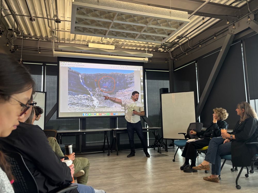 A man in front of a small audience, pointing at a photo of a landslide on a screen 