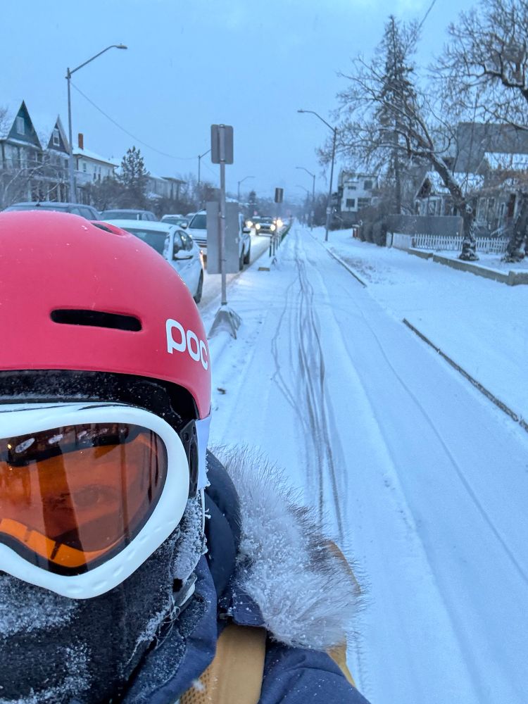 A woman wearing a sky helmet and goggles. In the background, there is a bike lane with a snow cover cut by some bike tire marks. 