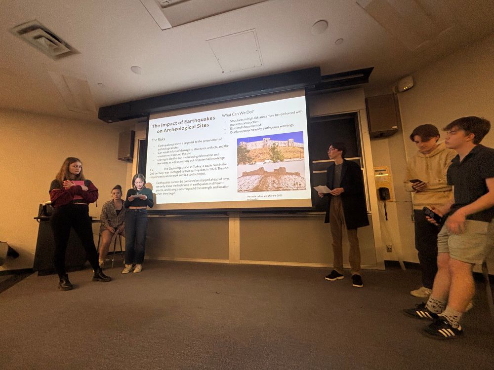 Students making a presentation on a a stage. The slide on the screen reads: 
The impacts of earthquakes on archaeological sites.