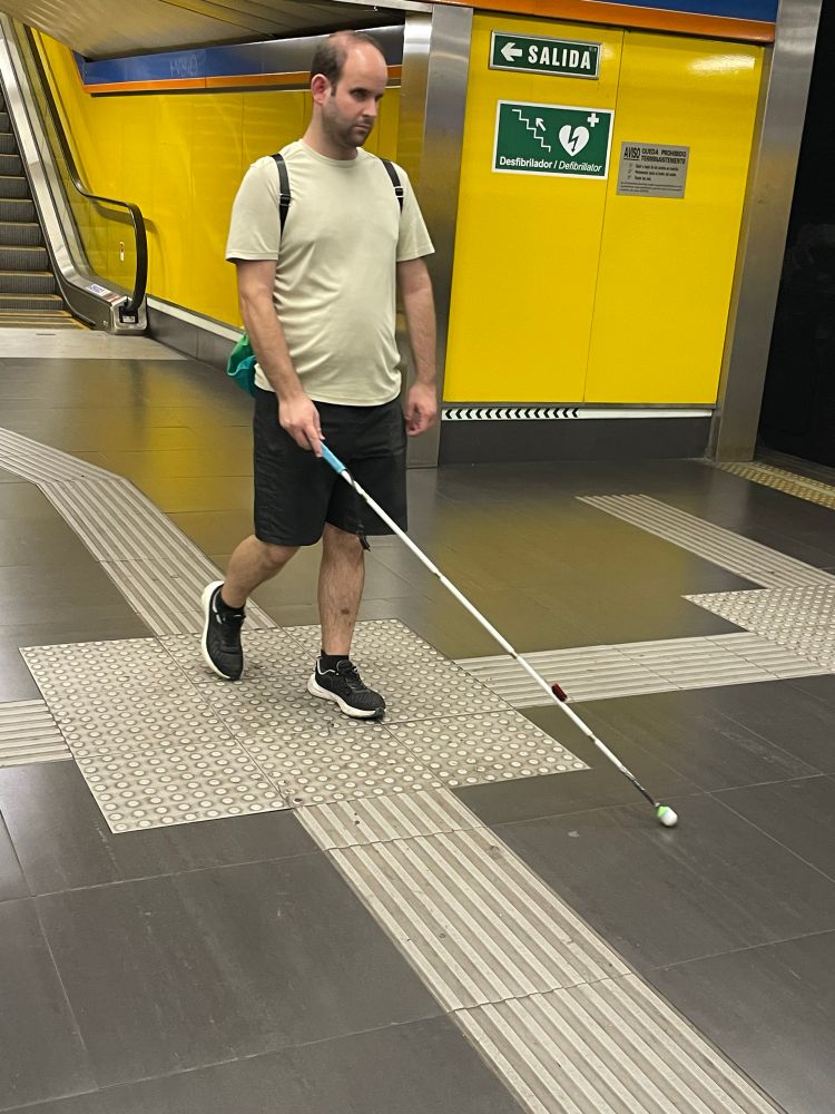 Dani caminando en una estación de la red de metro de Madrid. Lleva una camiseta beige, pantalones cortos negros, mochila y zapatillas deportivas negras. Utiliza un bastón blanco para personas con discapacidad visual y sigue el camino podotáctil en el suelo, diseñado para guiar a personas ciegas o con baja visión. Al fondo, las paredes son de color amarillo y hay señales que indican la salida y la ubicación de un desfibrilador. También se observa una escalera mecánica a la izquierda.
