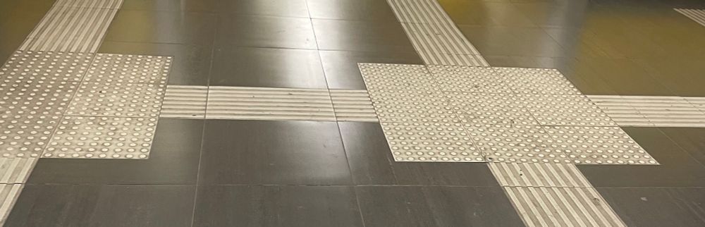 Piso de estación de metro con guías podotáctiles. Se observan franjas y cuadros en relieve sobre el suelo, diseñados para ayudar a personas con discapacidad visual a orientarse y desplazarse de manera segura. Las líneas rectas indican dirección, mientras que los cuadros con puntos en relieve marcan zonas de cruce o advertencia. A los costados, cerca del borde del andén, hay una franja amarilla también en relieve que señala el límite seguro antes de la vía del tren. Las paredes al fondo son de color amarillo brillante.
