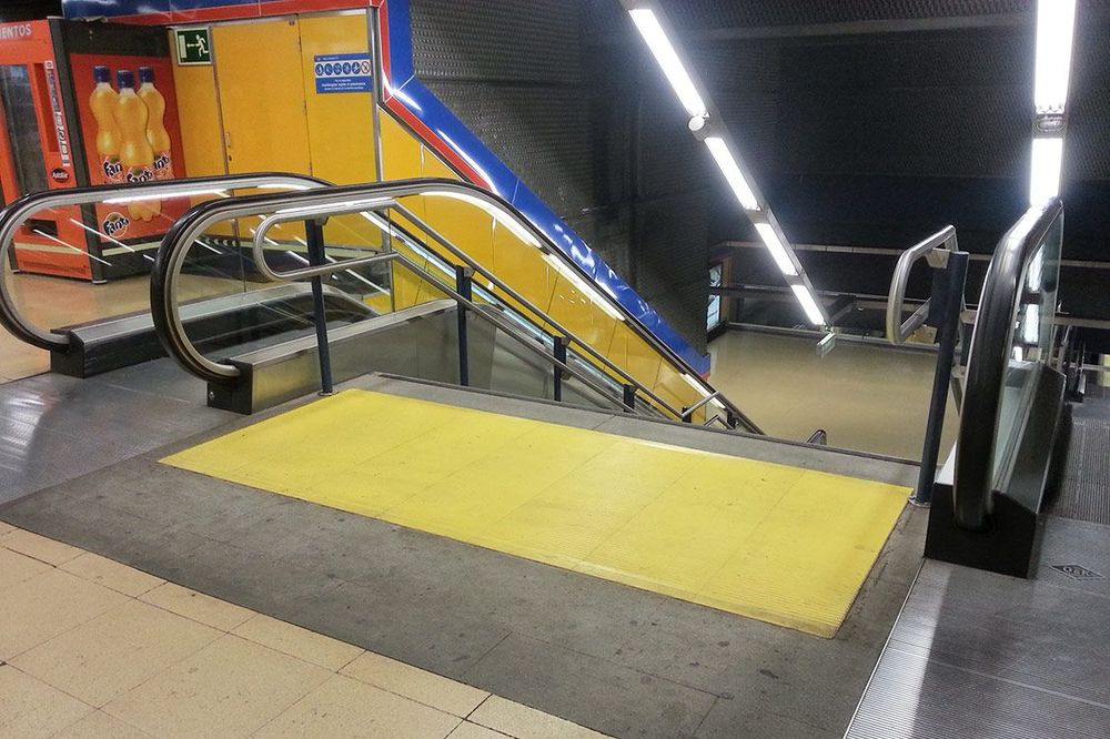 Escalera manual en una estación de la red de metro de Madrid. La escalera desciende en línea recta y está flanqueada por barandillas metálicas a ambos lados. Justo antes de comenzar los escalones, en el suelo, hay una franja podotáctil de color amarillo. Este podotáctil es rectangular, ocupa todo el ancho de la escalera y está compuesto por placas con relieve. El relieve consiste en líneas paralelas en sentido transversal a la marcha, diseñadas para ser detectadas fácilmente con el bastón o al pisar, alertando a las personas con discapacidad visual de la presencia de la escalera. El contraste del amarillo con el suelo gris facilita su identificación visual.
