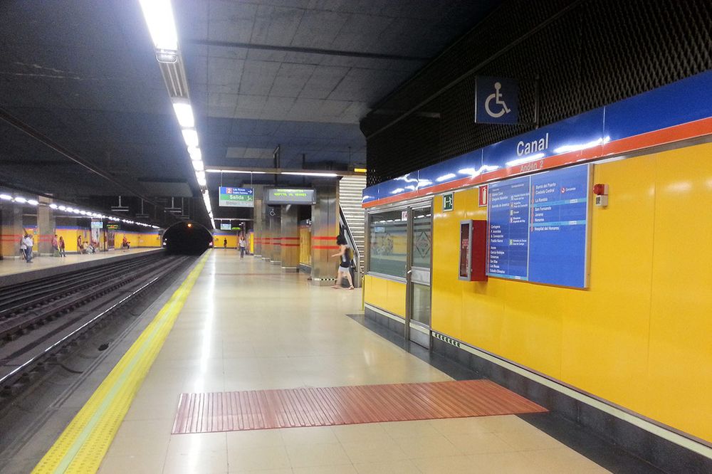 Andén de la estación de metro Canal. La imagen muestra un andén amplio y moderno, con paredes amarillas y detalles en azul y naranja. En la parte superior derecha se ve el cartel con el nombre de la estación: "Canal", y debajo indica "Andén 2".

En cuanto a las guías podotáctiles, se observan dos tipos: una franja podotáctil amarilla, que recorre el borde del andén junto a las vías, diseñada como guía de posicionamiento para indicar la zona segura donde esperar el tren y evitar caídas. Además, hay una superficie podotáctil roja, de forma rectangular, situada perpendicularmente a la franja amarilla, justo delante de la puerta de acceso al tren, que sirve para indicar el punto de parada y acceso al vagón.

No se observa ninguna guía podotáctil longitudinal a lo largo del andén, es decir, no hay una guía que permita a las personas con discapacidad visual orientarse caminando por el andén, más allá de las zonas de posicionamiento para abordar el metro. La estación está bien iluminada y cuenta con señalización clara, pero la accesibilidad podotáctil se limita a las zonas de espera y acceso al tren.